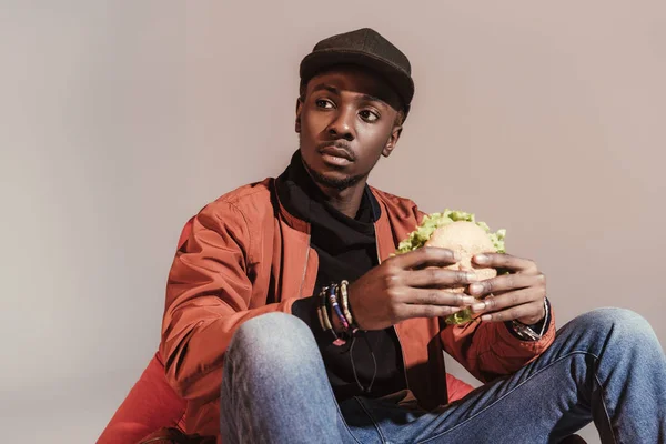 Fiatal Afroamerikai Férfi Eszik Hamburgert Keres Elszigetelt Szürke — Stock Fotó