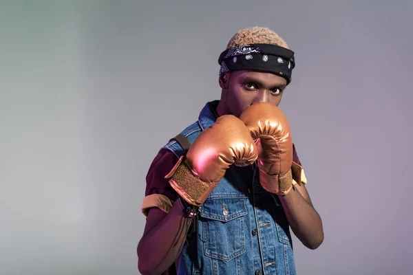 stylish young african american man in golden boxing gloves looking at camera isolated on grey