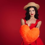 Attractive girl in red dress showing heart shaped pillow isolated on red