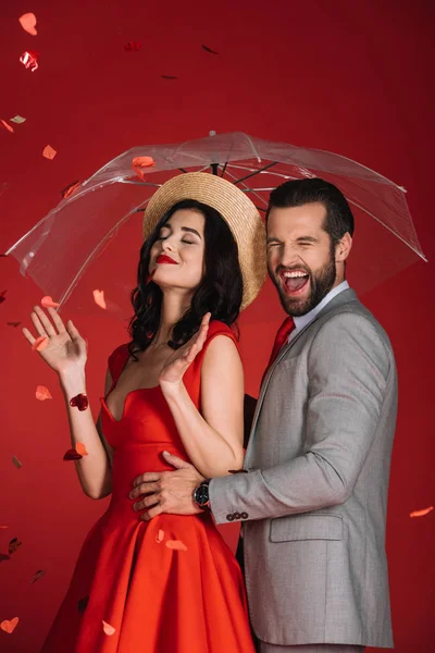 laughing couple with umbrella under falling confetti isolated on red