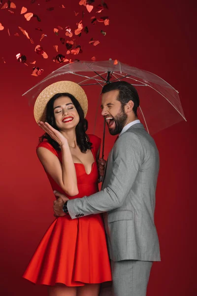 laughing couple hiding under umbrella from falling confetti isolated on red