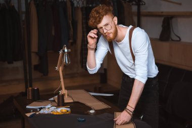 genç erkek moda tasarımcısı dikiş desenleri ve kumaş ile çalışırken kameraya bakarak gözlük