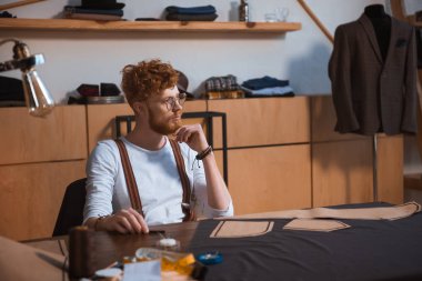düşünceli erkek moda tasarımcısı uzakta dikiş desenleri ve kumaş ile masada otururken seyir gözlük