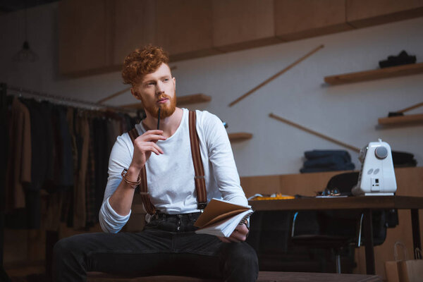 pensive young male fashion designer looking away while taking notes in notebook at workplace
