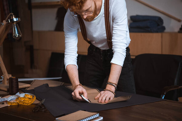 concentrated young male fashion designer in eyeglasses working with sewing pattern in workshop 