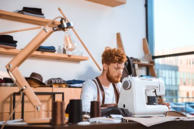 Sakallı genç moda tasarımcı atölyesinde dikiş makinesi ile çalışma