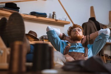 seçici odak önlük ve gözlük elle işyerinde başının arkasında oturan genç erkek moda tasarımcısı