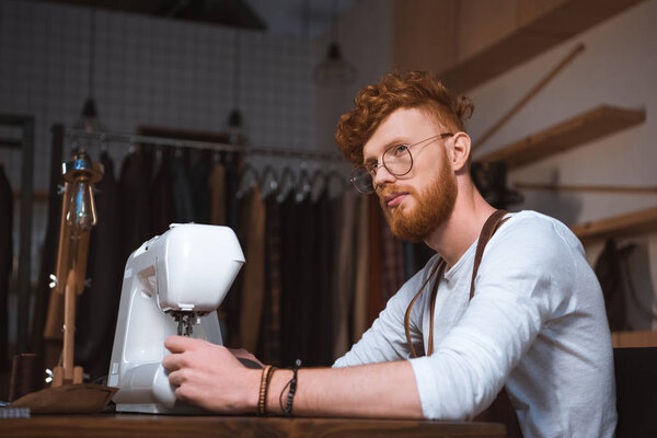 задумчивый молодой модельер работает с швейной машинкой и смотрит в сторону мастерской
