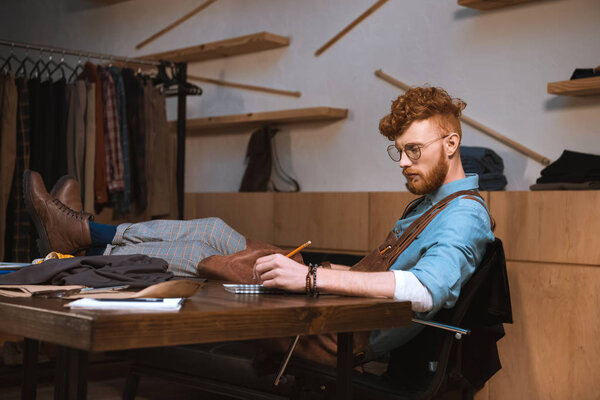 young male fashion designer in eyeglasses taking notes at workplace