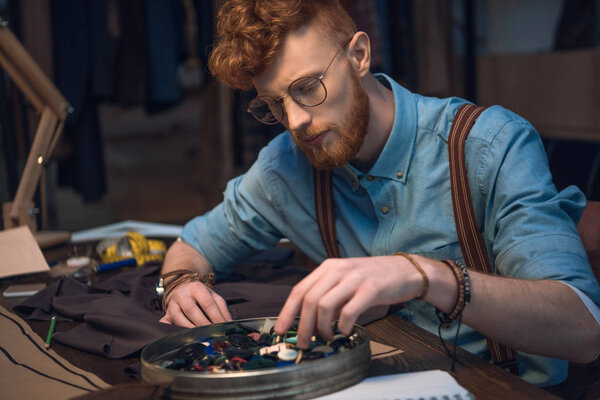 young male fashion designer in eyeglasses working with colorful buttons and fabric at workshop