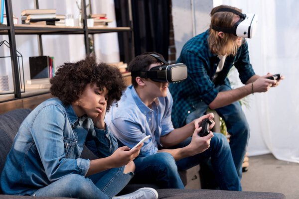 friends playing video game with virtual reality headsets, african american girl sitting with smartphone
