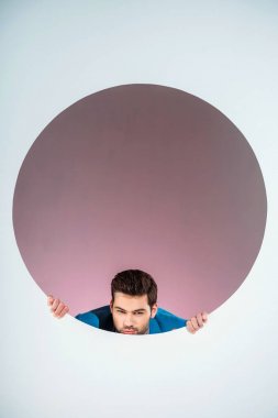 stylish bearded young man looking at camera through hole on grey  clipart