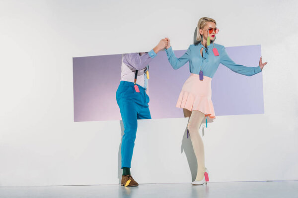 stylish young couple with colorful tags on clothes walking through opening on grey