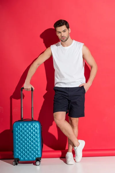 handsome young man standing with suitcase and looking at camera on pink