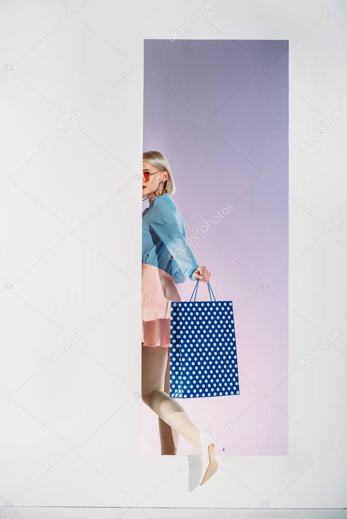 stylish young woman with shopping bag stepping through aperture on grey