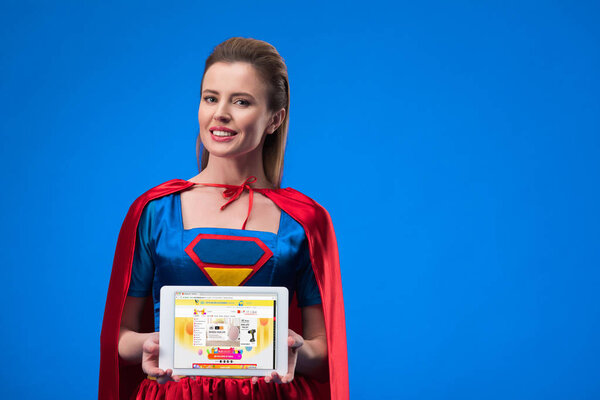 portrait of smiling woman in superhero costume showing tablet isolated on blue