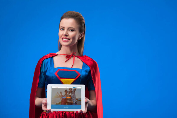 portrait of cheerful woman in superhero costume showing tablet isolated on blue