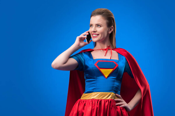 portrait of smiling woman in superhero costume talking on smartphone isolated on blue