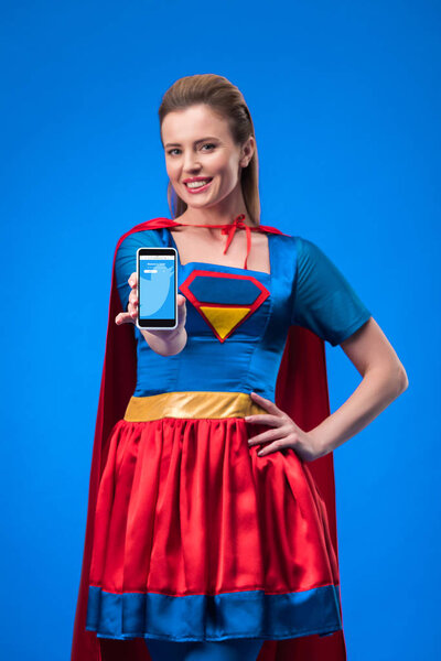 portrait of smiling woman in superhero costume showing smartphone isolated on blue