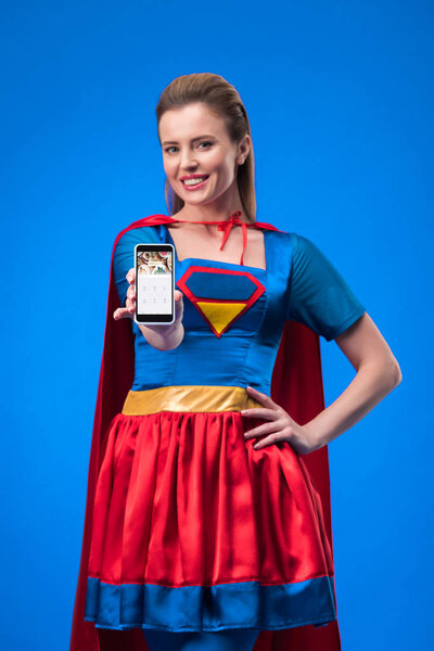 portrait of smiling woman in superhero costume showing smartphone isolated on blue