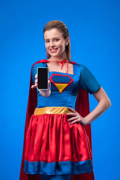 portrait of smiling woman in superhero costume showing smartphone with blank screen isolated on blue