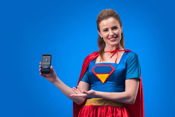 Portrait of smiling woman in superhero costume showing smartphone with tumblr app isolated on blue
