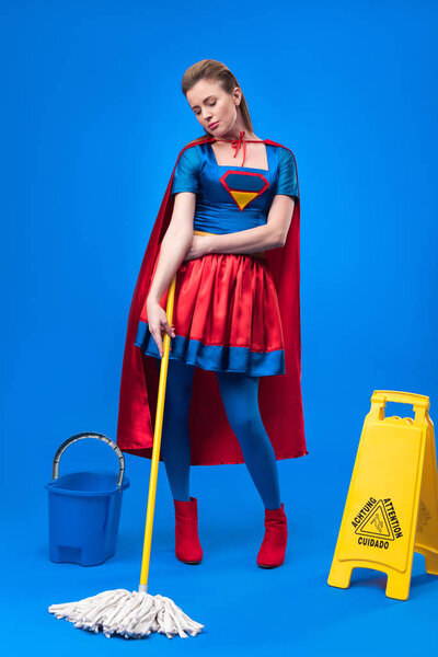 attractive woman in superhero costume with caution sign, bucket and mop for cleaning isolated on blue