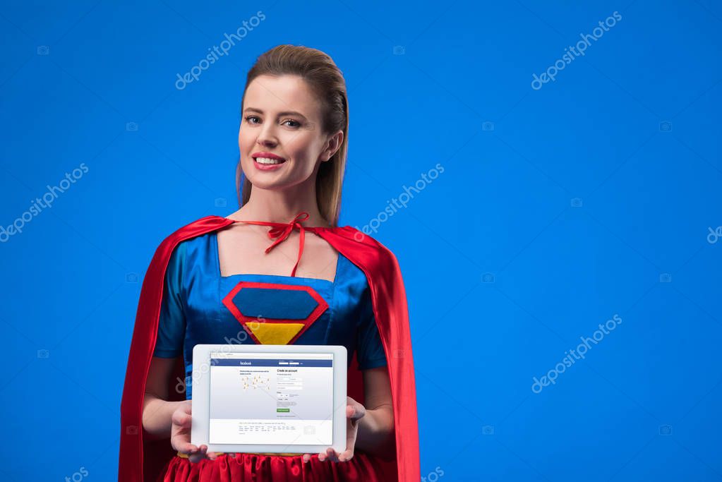 Portrait of cheerful woman in superhero costume showing tablet isolated on blue