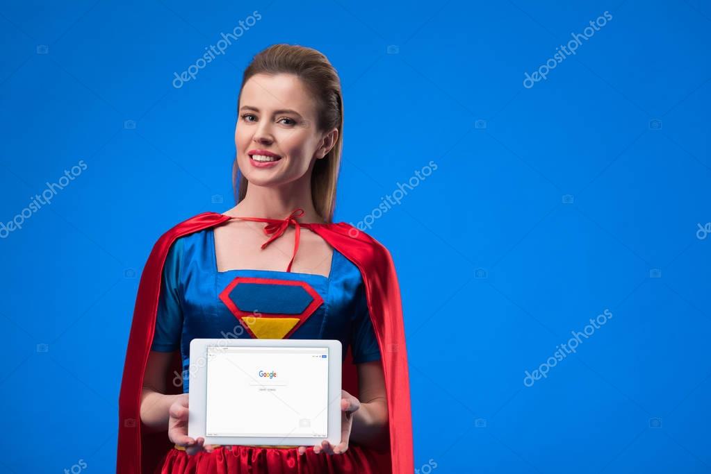 Portrait of cheerful woman in superhero costume showing tablet isolated on blue