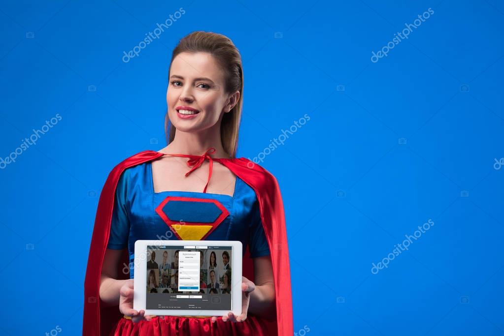Portrait of cheerful woman in superhero costume showing tablet isolated on blue