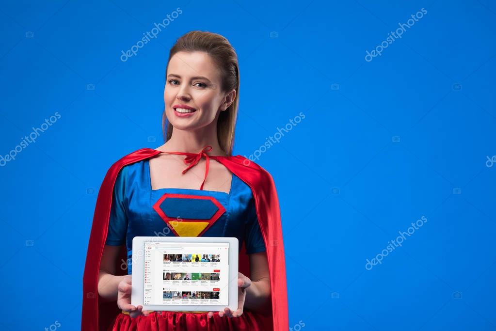 Portrait of cheerful woman in superhero costume showing tablet isolated on blue
