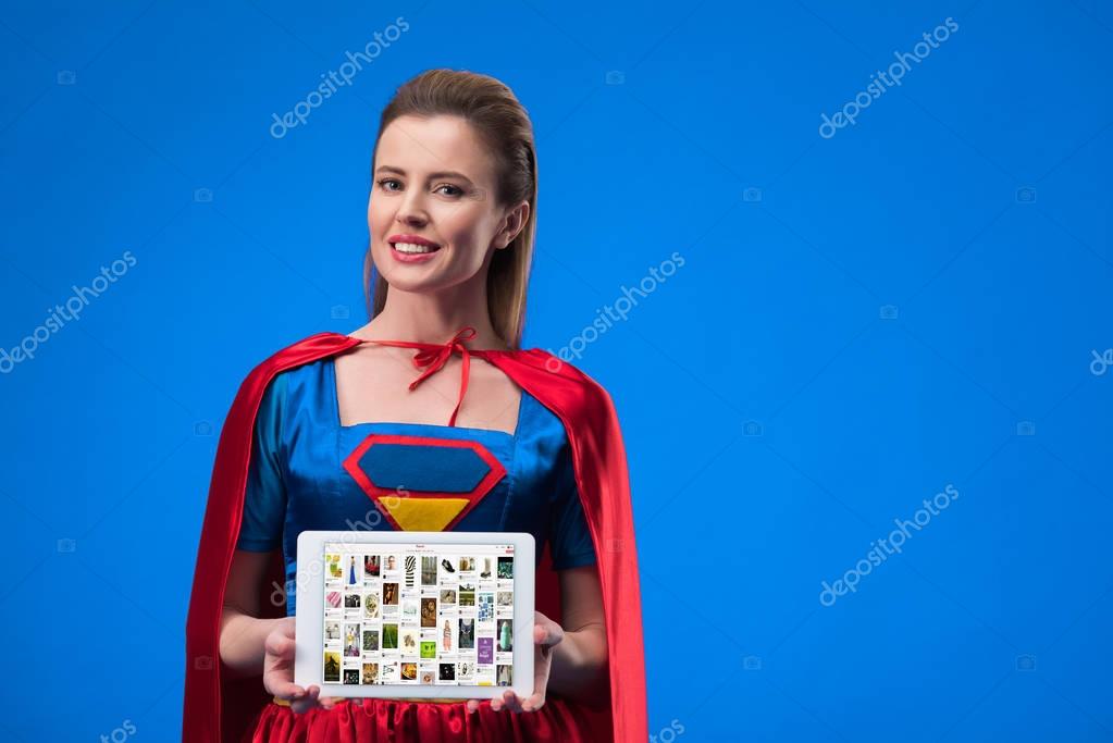 Portrait of cheerful woman in superhero costume showing tablet isolated on blue