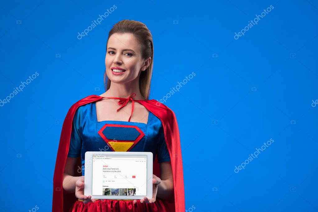Portrait of cheerful woman in superhero costume showing tablet isolated on blue