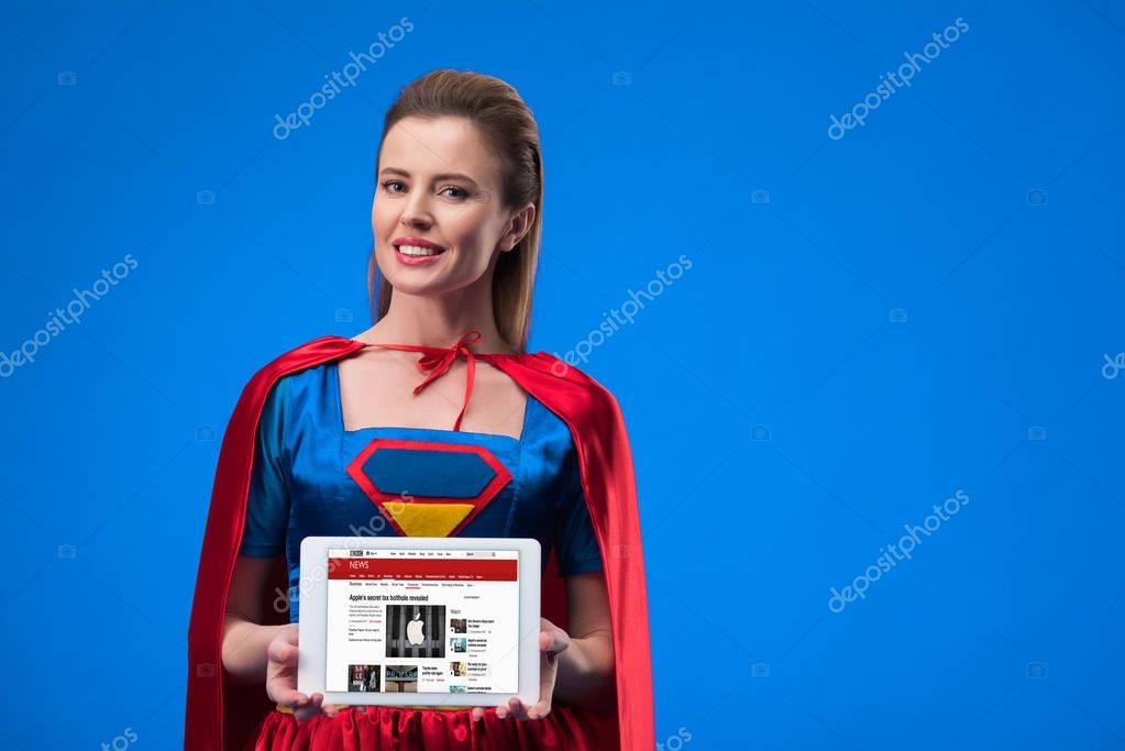 Portrait of cheerful woman in superhero costume showing tablet isolated on blue