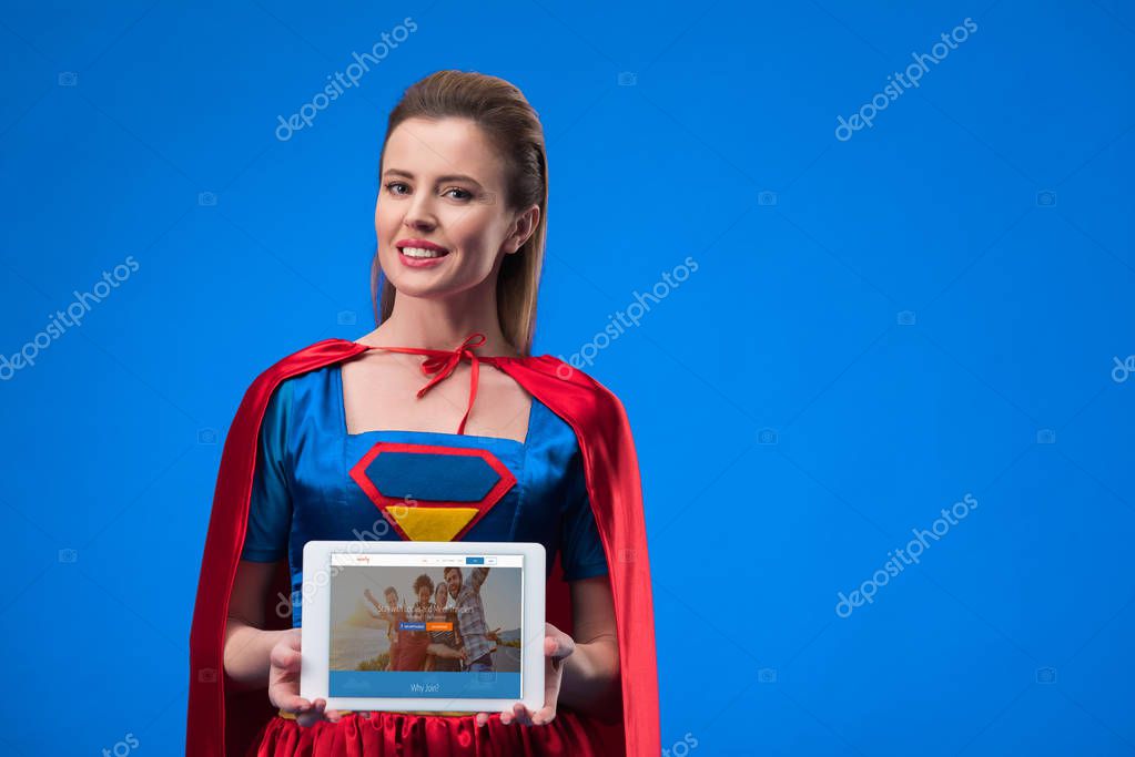 Portrait of cheerful woman in superhero costume showing tablet isolated on blue