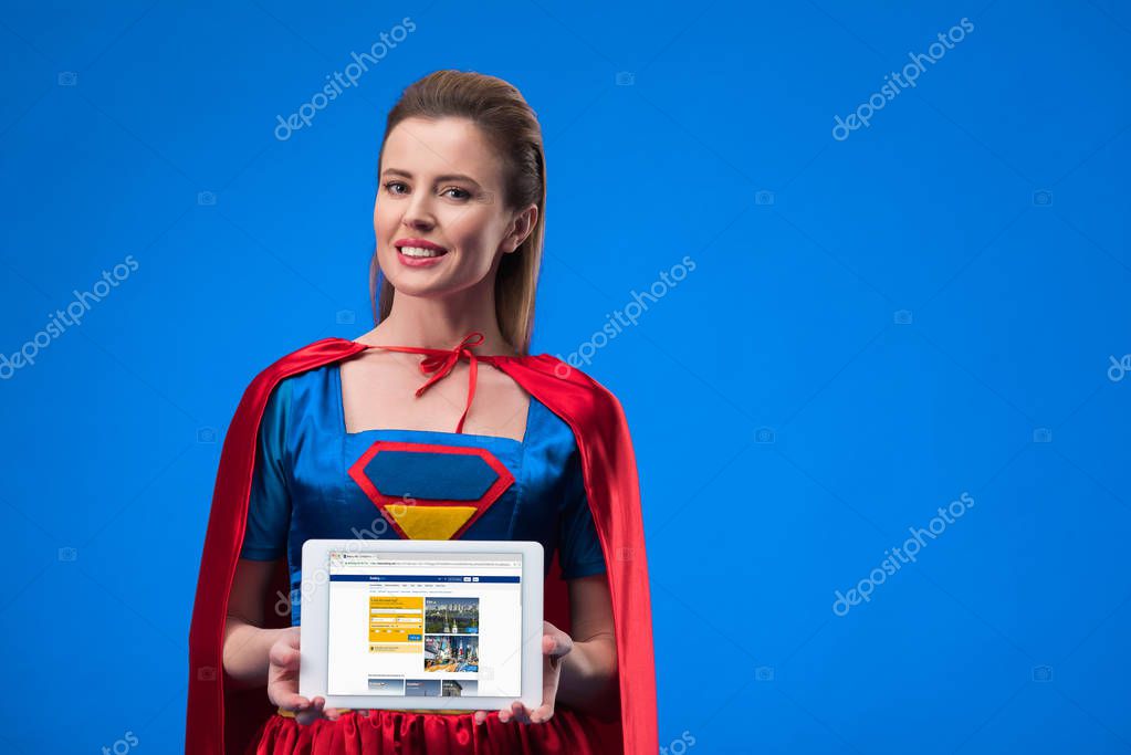 Portrait of smiling woman in superhero costume showing tablet isolated on blue