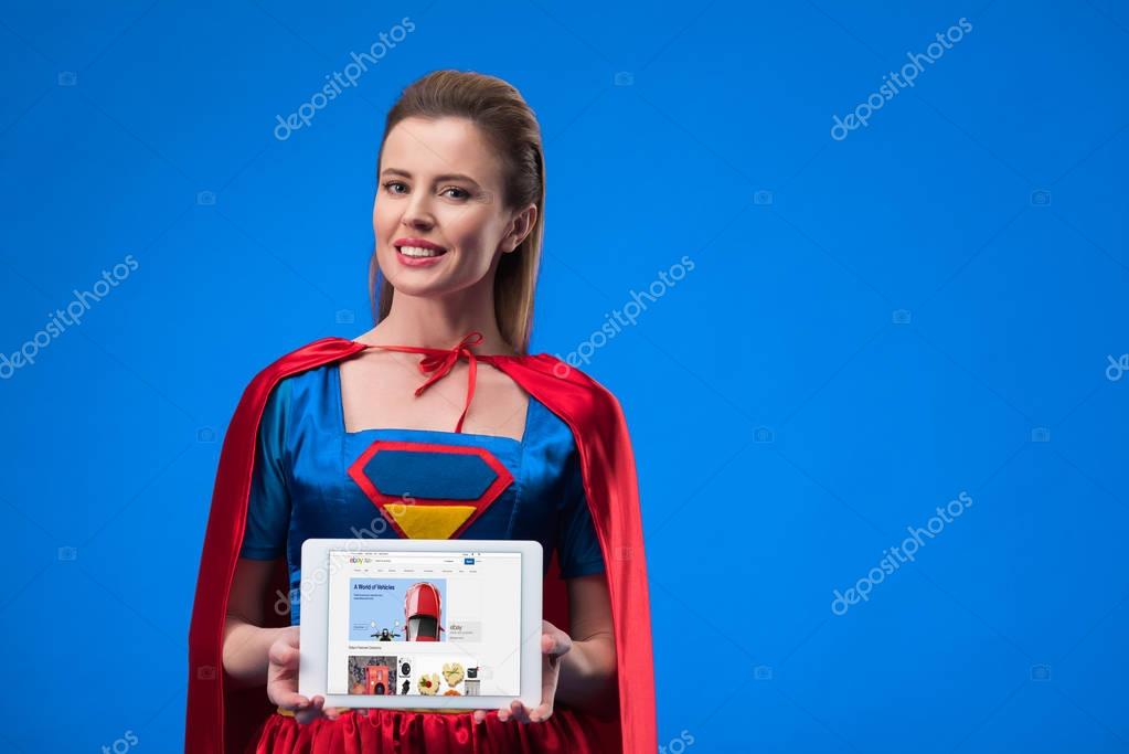 Portrait of smiling woman in superhero costume showing tablet isolated on blue