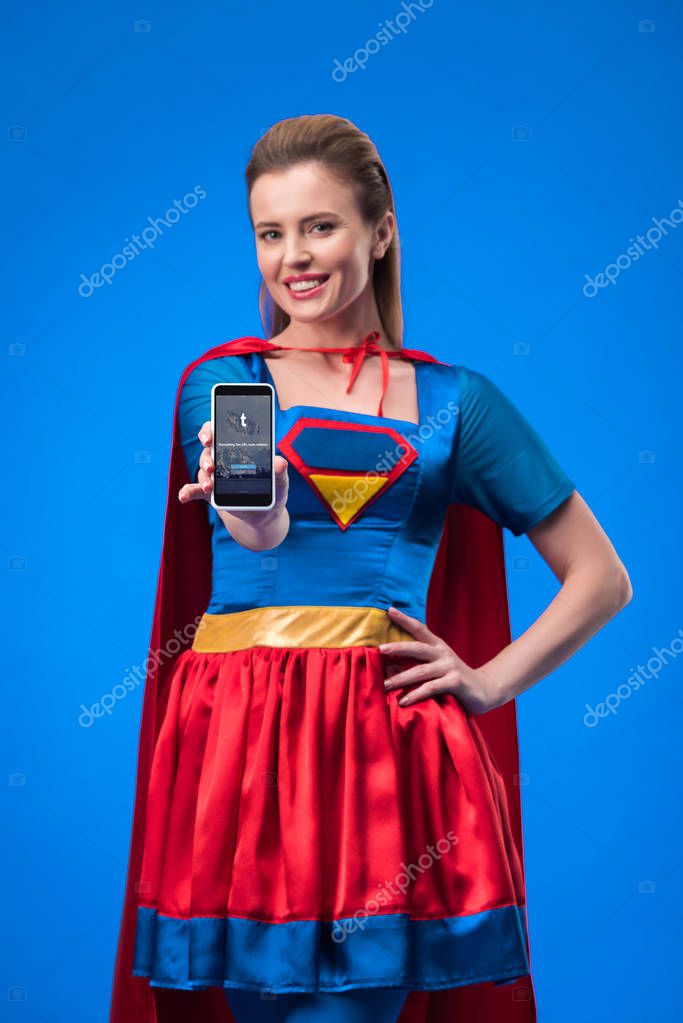 Portrait of smiling woman in superhero costume showing smartphone with tumblr app isolated on blue