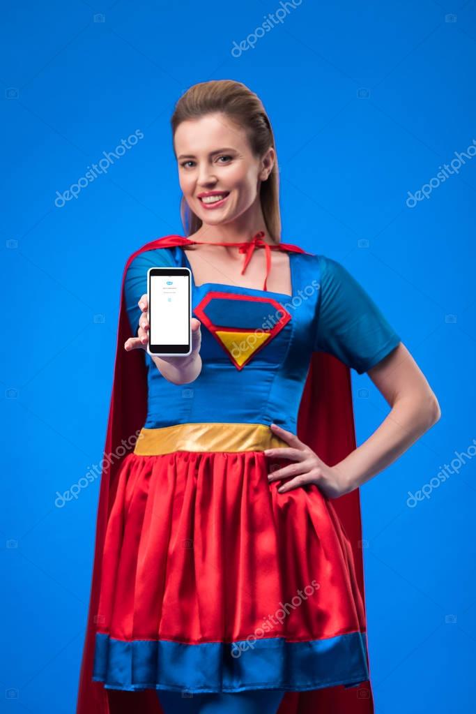 Portrait of smiling woman in superhero costume showing smartphone isolated on blue