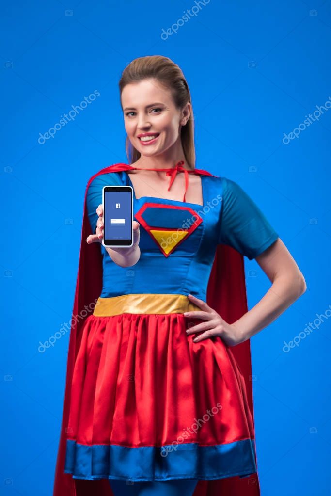 Portrait of smiling woman in superhero costume showing smartphone isolated on blue