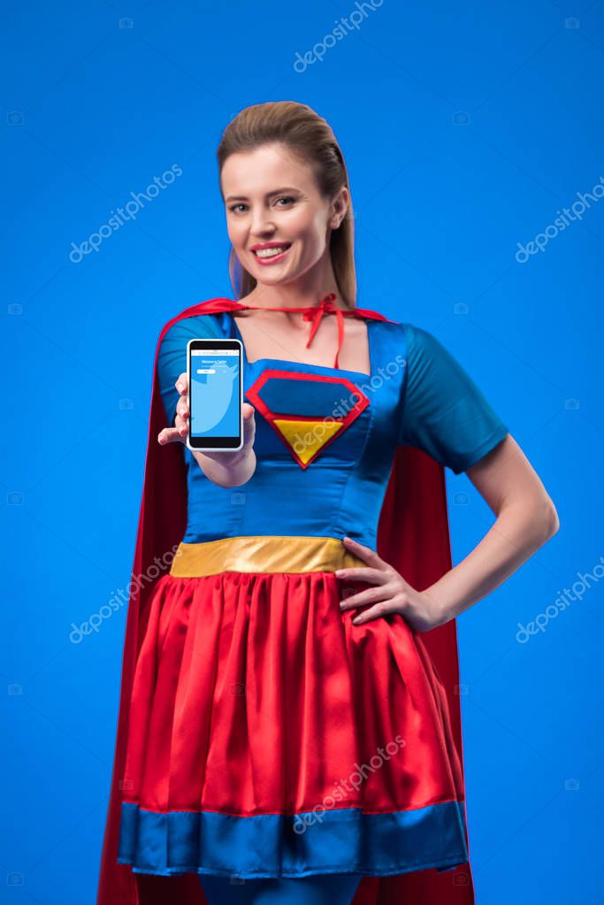 Portrait of smiling woman in superhero costume showing smartphone isolated on blue