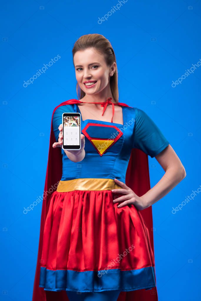 Portrait of smiling woman in superhero costume showing smartphone isolated on blue