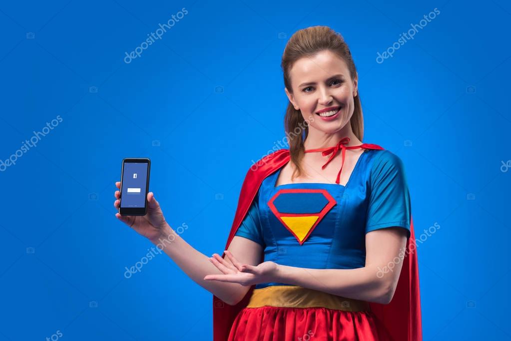 Portrait of smiling woman in superhero costume showing smartphone with facebook app isolated on blue