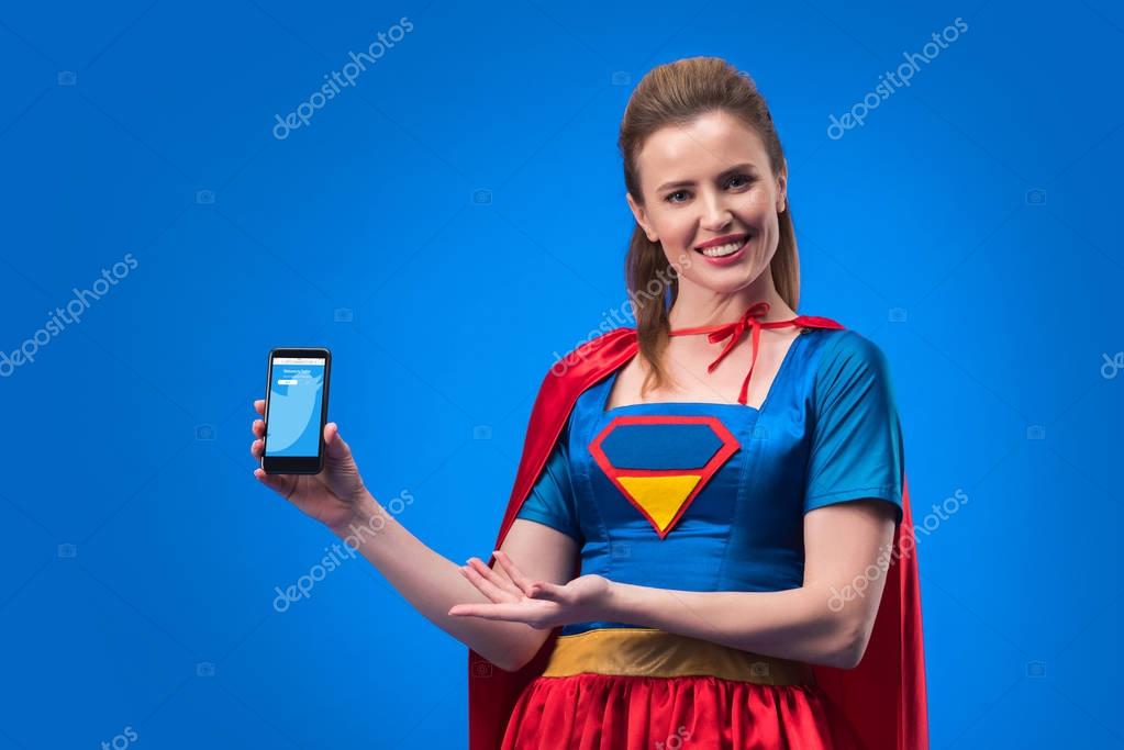 Portrait of smiling woman in superhero costume showing smartphone isolated on blue