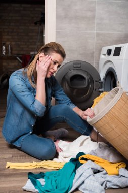 emotional housewife looking at clothing before putting it into washing machine at home clipart