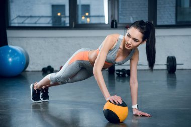 young sporty woman doing push ups with one hand on ball at gym clipart