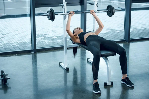 年轻的运动女子举重杠铃躺在健身房的长凳上 — 图库照片