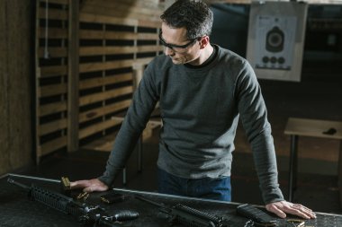 handsome man leaning on table with weapons in shooting range clipart