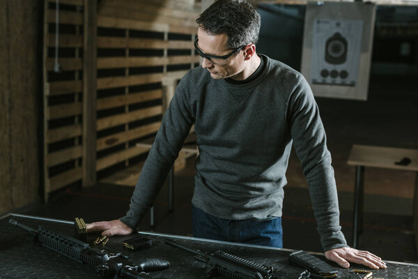 handsome man leaning on table with weapons in shooting range