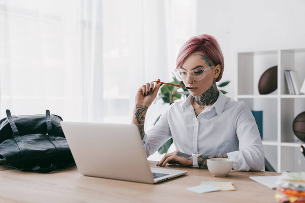 young tattooed businesswoman holding pencil and using laptop at workplace